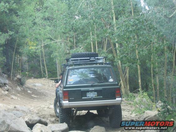 Dark green 1996 Jeep Cherokee with a roof rack and spare tire, driving on a rocky, wooded trail