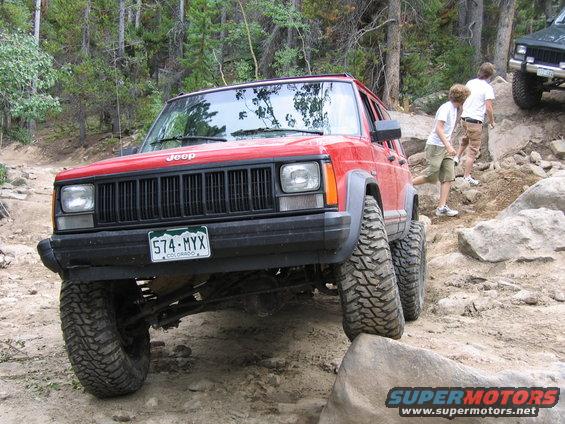 Red 1996 Jeep Cherokee navigating a rocky trail with two people walking behind it. Another green Jeep is visible further up the trail
