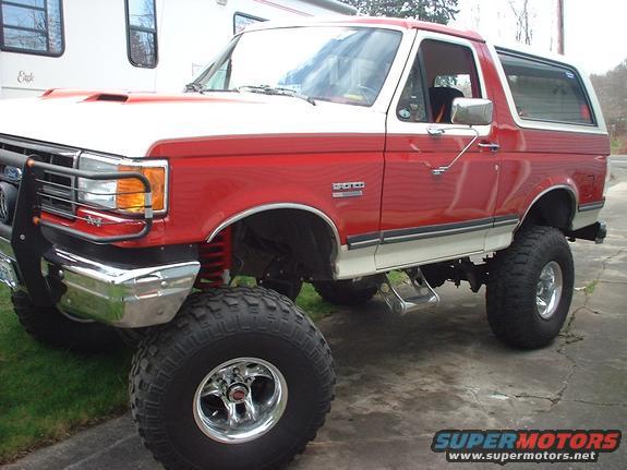 Red and white lifted Ford Bronco with large tires, chrome bumper, and brush guard