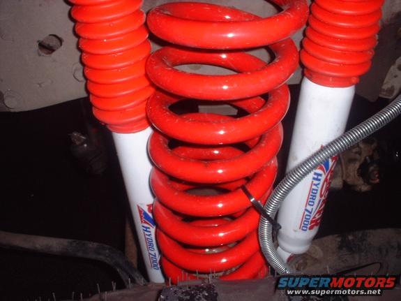 Close-up of red coil springs and white shock absorbers with "HYDRO 7000" text. Bottom has "SUPERMOTORS" logo