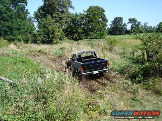 Black 1978 Ford Bronco driving through a muddy field, rear view
