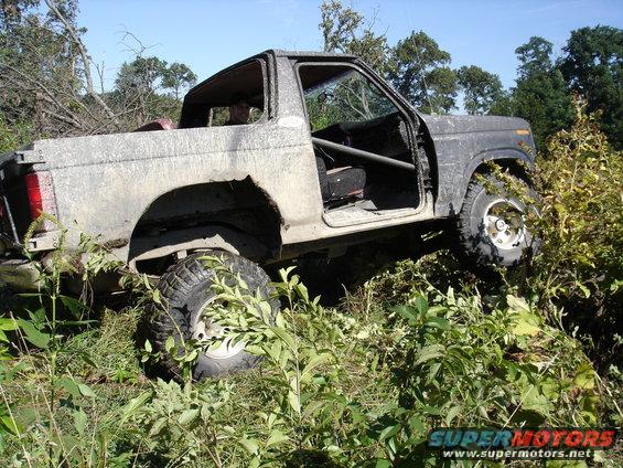 1979 Ford Bronco with roll cage, doors removed, covered in mud, driving through brush
