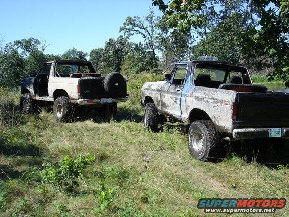 Two Ford Broncos parked in a grassy field, one with a spare tire mounted on the rear. The Bronco on the right has a blue and white paint scheme, while the one on the left is mostly white