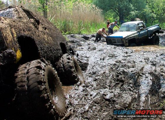 Two trucks stuck deep in mud, one on its side, with people trying to push the other