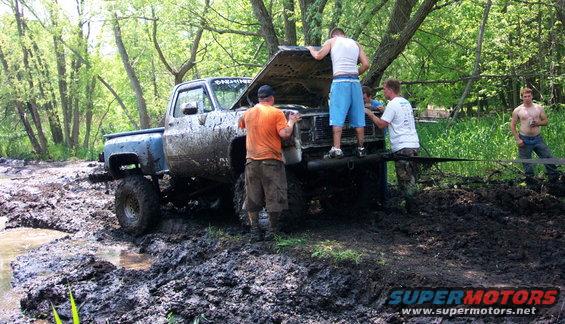 Mud-covered pickup truck with hood open, people gathered around the engine, stuck in a muddy wooded area