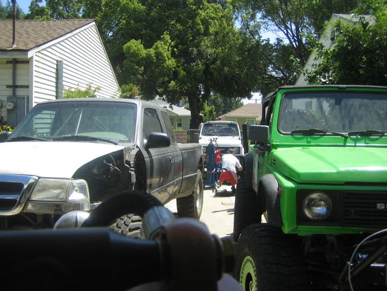 Green Suzuki Samurai front view with tube bumper. A black pickup truck and white vehicle are also visible