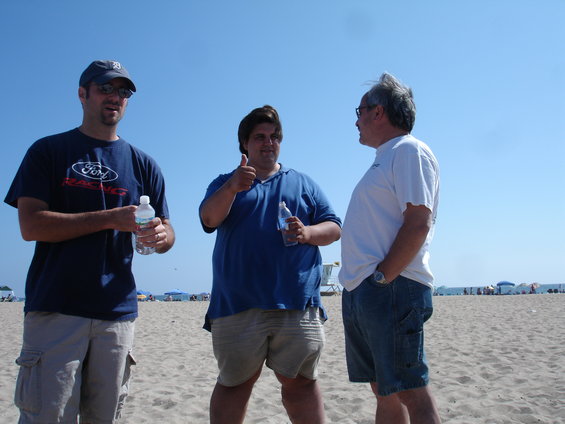 Three men stand on a sandy beach under a clear blue sky. The man on the left wears a dark blue t-shirt with a Ford logo