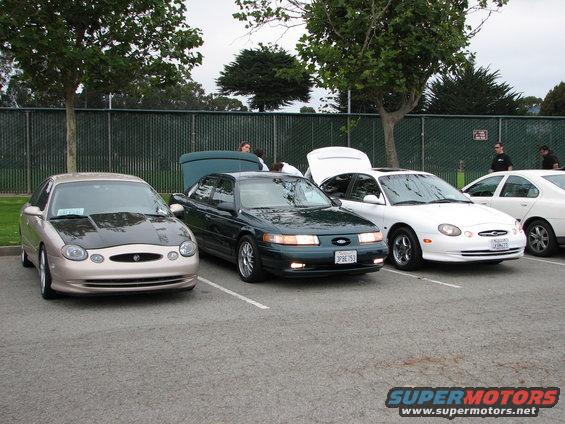 Three sedans parked in a lot, one tan with a black hood, one dark green, and one white