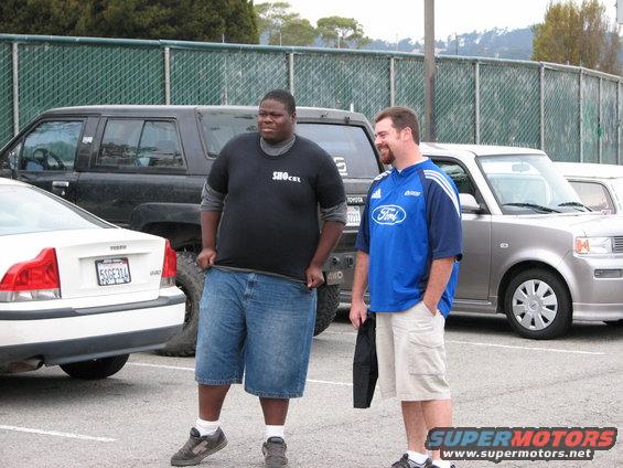 Two men stand in a parking lot next to a white Volvo and a silver Toyota