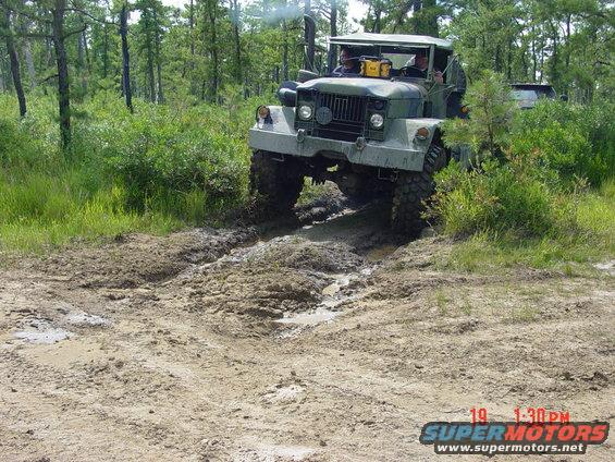 Military truck with large tires driving through a muddy wooded area. Another vehicle is partially visible behind it