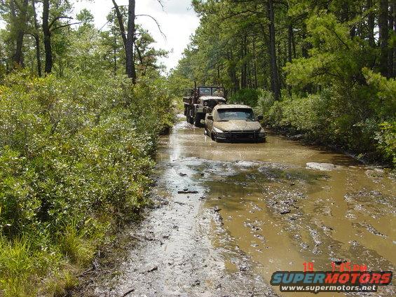 Two trucks driving through a muddy, wooded area
