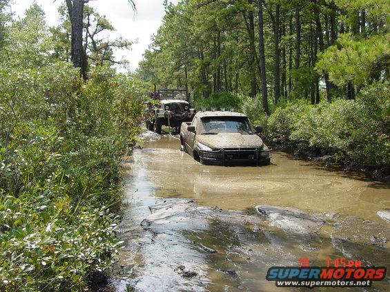 Mud-covered pickup truck stuck in a muddy water trail, front view. A military-style vehicle follows behind