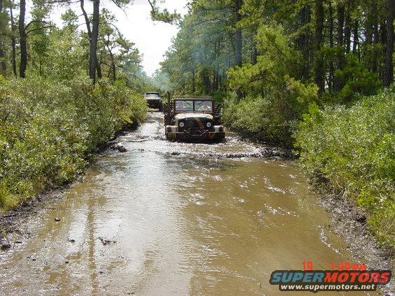 Two old military trucks driving through a muddy, wooded trail