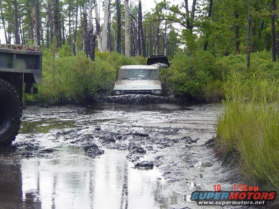 Off-road vehicles stuck in a muddy forest path. A large truck is on the left, and two SUVs are in the mud ahead