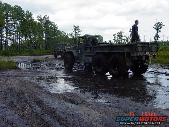 Two olive drab military trucks stuck in mud, one with a man standing in its bed
