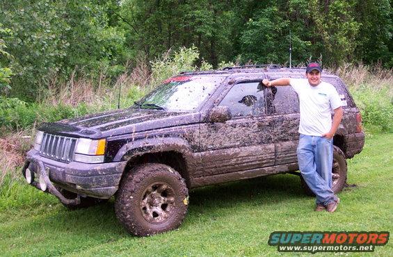 Guy leaning on a muddy Jeep Grand Cherokee parked on grass with trees in the background
