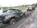1990 Ford Bronco - A black pickup truck is stuck in deep mud and being towed by a vintage military truck on a dirt road.