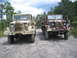 1990 Ford Bronco - Two military trucks are parked side-by-side on a dirt road with trees in the background.