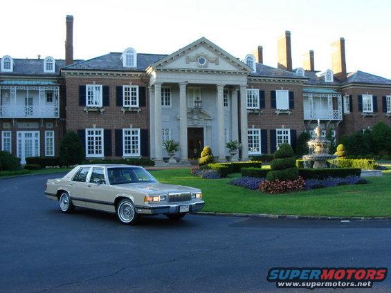 Tan 1990 Mercury Grand Marquis sedan parked on a dark asphalt driveway in front of a large brick mansion with a fountain
