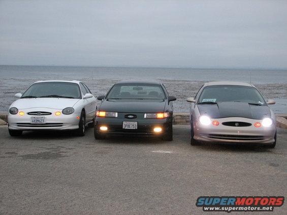 Three sedans, a white Ford Taurus, a dark Ford Taurus, and a silver Ford Taurus with a black hood, parked in front of the ocean