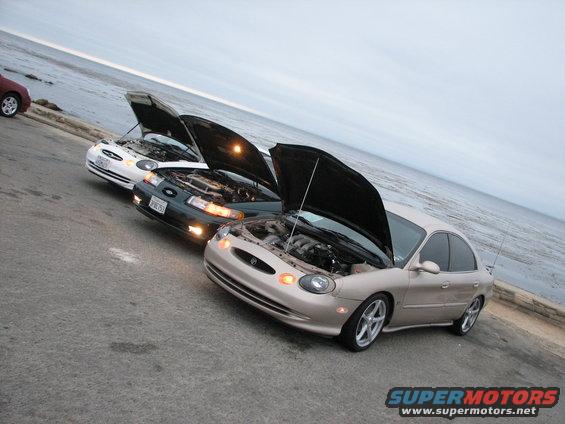 Tan sedan with hood open, parked next to two other sedans by the water. License plate visible