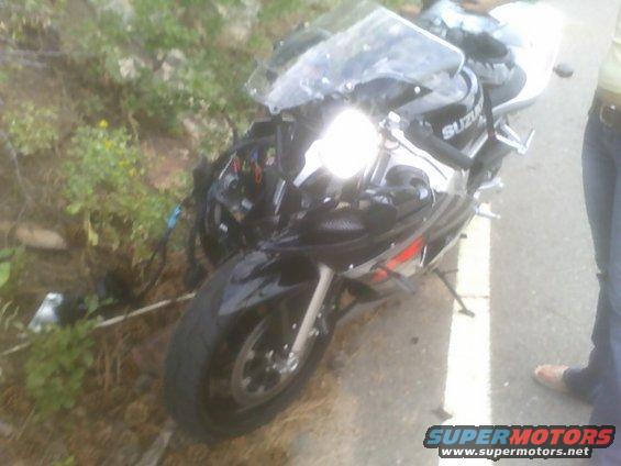 Black Suzuki sportbike with damaged front end, off the road next to some bushes. A person stands nearby