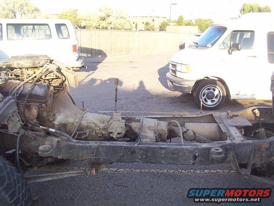 Chassis of a vehicle with engine and transmission visible. Two white vans are parked in the background