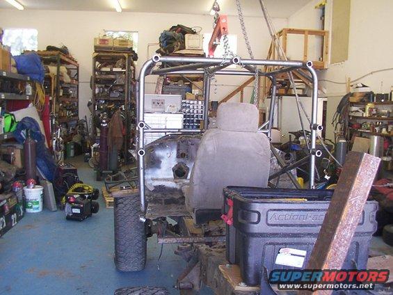 Vehicle chassis with roll cage, gray seat, and exposed engine bay, suspended in a garage