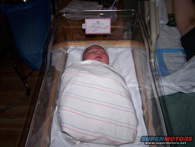 Newborn baby wrapped in a striped blanket, sleeping in a clear hospital bassinet. A pink card is attached to the side