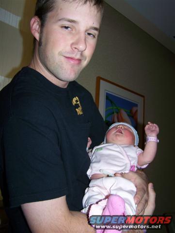 Man in black t-shirt holding a baby wearing a pink hat and bracelet