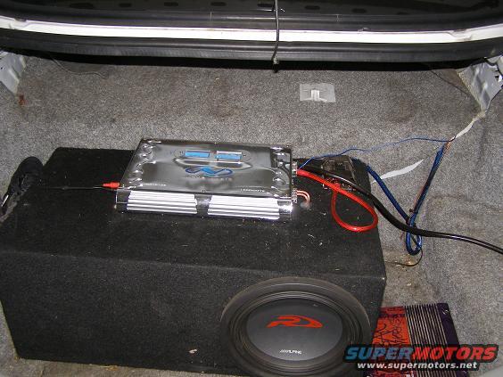 Car trunk interior with a subwoofer, amplifier, and wiring. A black shoe is visible on the left