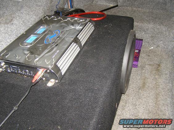 Car audio amplifier with blue lights and red/black RCA cables connected to a subwoofer with a purple component visible