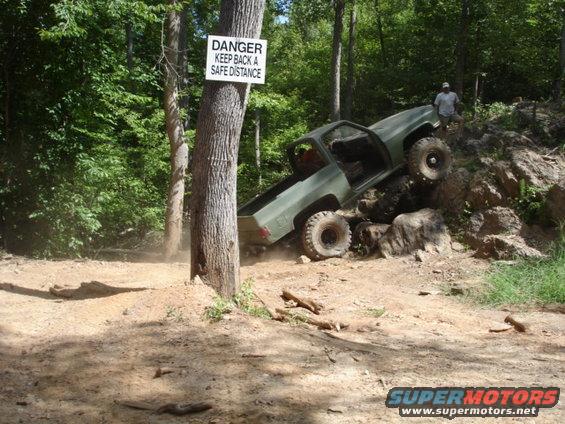 Off-road truck climbing a rocky incline with a 'DANGER KEEP BACK A SAFE DISTANCE' sign on a tree