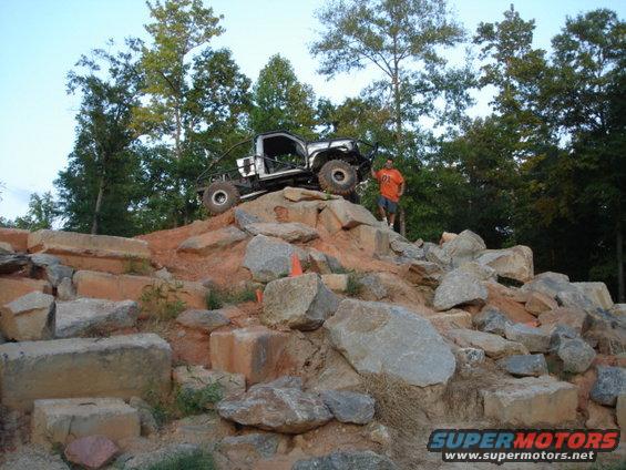 Modified off-road truck with roll cage climbing large rocks. Man stands on rock next to vehicle