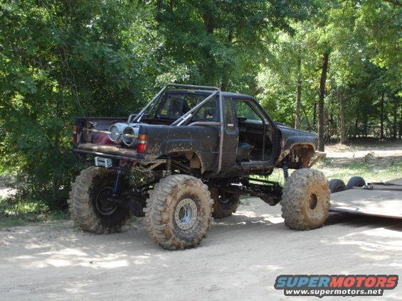 muddy pickup truck with huge tires, roll cage, and exposed doors, parked on dirt near trees