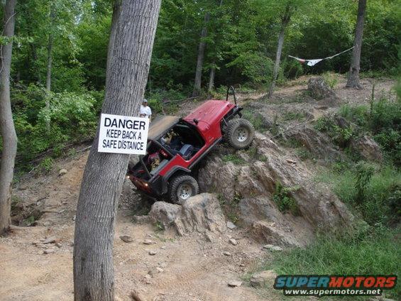 Red Jeep Wrangler climbing a rocky hill with a danger sign on a tree