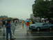 1978 Ford Mustang II - A blue coupe is parked on a wet street during a car show, with people holding umbrellas.
