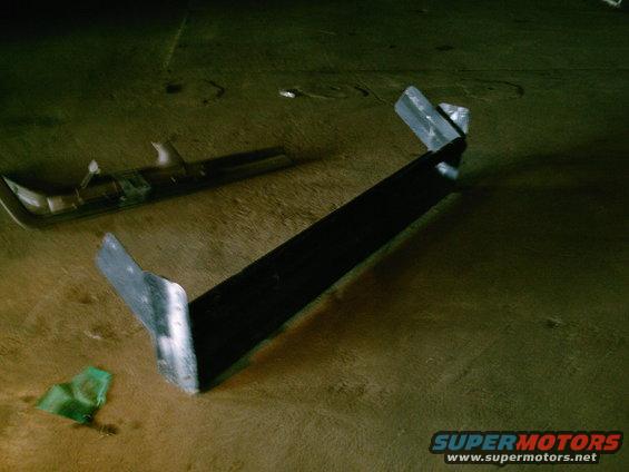 Black metal vehicle part with silver mounting brackets on a wooden surface. Another chrome-colored part is visible in the background