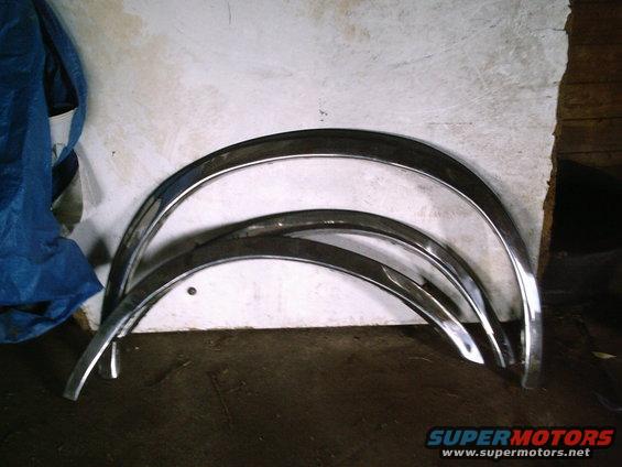 Three chrome fender trim pieces with a blue tarp and wooden wall in the background