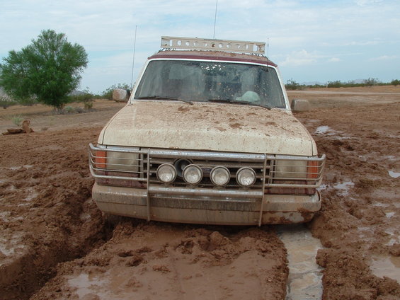 Mud-covered white SUV with a roof rack stuck in deep mud, front view