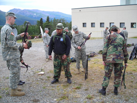 Soldiers in camouflage uniforms stand outdoors near a building, some holding rifles