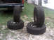 1996 Ford Bronco - Two pairs of off-road tires are stacked on gravel, with the rear of a dark green vehicle visible in the background.