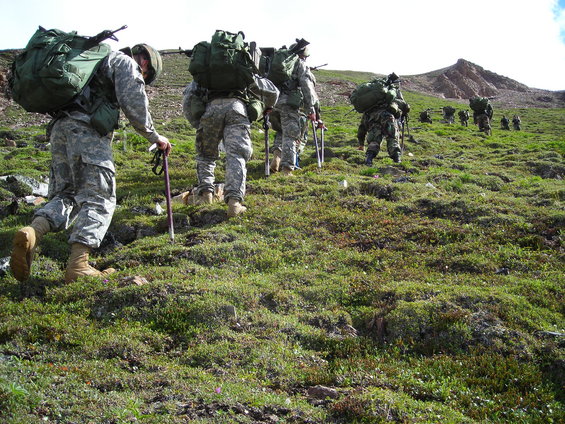 Soldiers with large backpacks hiking up a grassy, rocky hill