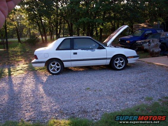 White coupe parked outside with hood up, trees in background. Blue SUV and another vehicle behind it