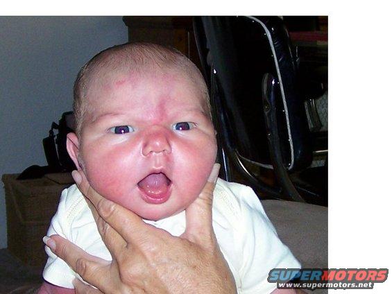 Baby with mouth open, held by a hand. Part of a black seat is visible
