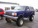 1989 Ford Bronco - This lifted blue Ford Bronco is parked next to a red SUV on a wet asphalt lot.