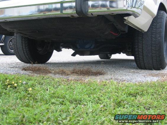 Underside of a light-colored sedan, showing rear axle, exhaust, and tires. A second vehicle is partially visible on the left