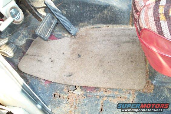 Floor mat and pedals in a vehicle, showing rust on the floor pan. Seat upholstery visible