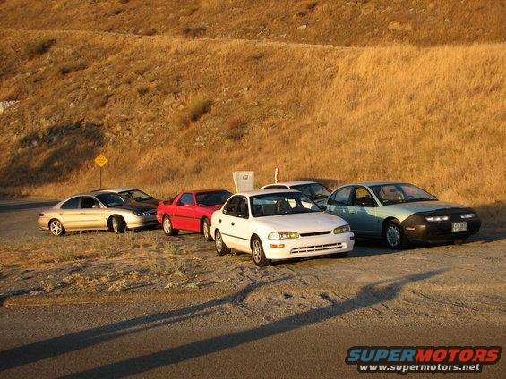 several cars parked on a dirt lot with a dry, grassy hill behind them