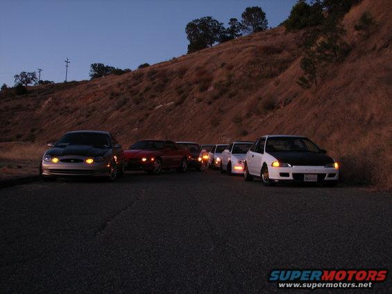 Several cars parked on a road with a hill in the background at dusk